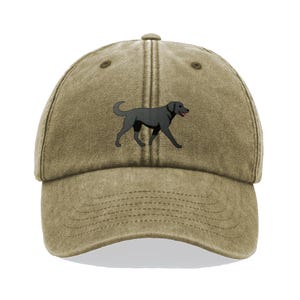 May include: A khaki-coloured baseball cap featuring an embroidered black Labrador Retriever. The dog is shown walking, with its tongue visible. The cap has a curved peak and an adjustable strap at the back.