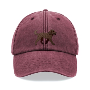May include: A maroon baseball cap featuring a brown dog graphic. The cap has a curved brim and an adjustable strap. The dog illustration is detailed with a textured appearance, adding visual interest to the hat.