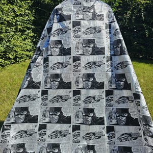 May include: A black and white comic book-style barber cape featuring panels with a masked character, a car, and speech bubbles. The cape is draped over a mannequin and is made of a shiny, reflective material.