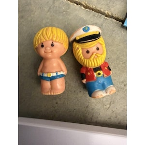 Może przedstawiać: Dwie małe plastikowe figurki. Jedna to blond dziecko w niebieskich szortach z białym paskiem. Druga to brodata postać w białej czapce kapitańskiej, czerwonej kurtce i niebieskich spodniach. Obie mają namalowane rysy twarzy i formowane włosy.