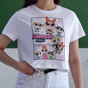 Puede incluir: Top corto blanco con un estampado gráfico de los personajes de dibujos animados de las Supernenas. El diseño incluye el logotipo del programa y varias imágenes de los personajes. La camiseta tiene mangas cortas y cuello redondo.