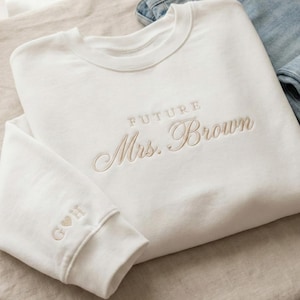 May include: A white sweatshirt with the embroidered text "FUTURE Mrs. Brown" in a delicate script. The sleeve cuff features initials "G & H" with a heart symbol. Light blue jeans are visible in the background. A cozy, casual bridal garment.