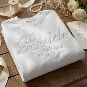 May include: A white sweatshirt with the word "Bride" embroidered in elegant script. Below the word "Bride" is a bow and the year "2026". The sweatshirt is folded on a wooden surface, with wedding-themed accessories in the background.