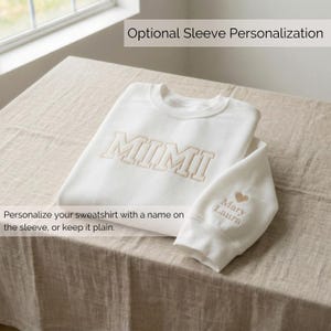 Op de afbeelding: Een witte sweatshirt met de naam "MIMI" in beige geborduurd over de borst. De mouw is gepersonaliseerd met de naam "Mary Laura" en een hart. De tekst "Optionele mouwpersonalisatie" en "Personaliseer je sweatshirt met een naam op de mouw, of houd het effen" zijn zichtbaar.
