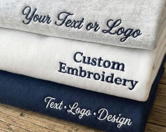 Op de afbeelding: Een stapel van drie gevouwen sweatshirts in lichtgrijs, wit en marineblauw. Het bovenste grijze sweatshirt heeft donkerblauw borduurwerk met de tekst Your Text or Logo. Het witte sweatshirt bevat de geborduurde tekst Custom Embroidery.