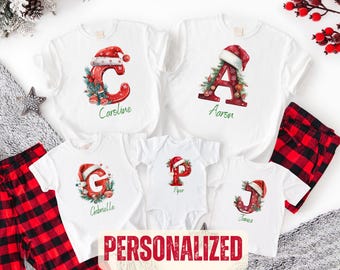 May include: A collection of white t-shirts and a baby bodysuit featuring red Christmas-themed letters C, A, G, P, and J, each adorned with a Santa hat and greenery. The names Caroline, Aaron, Gabrielle, Piper, and Jones are printed in green script.