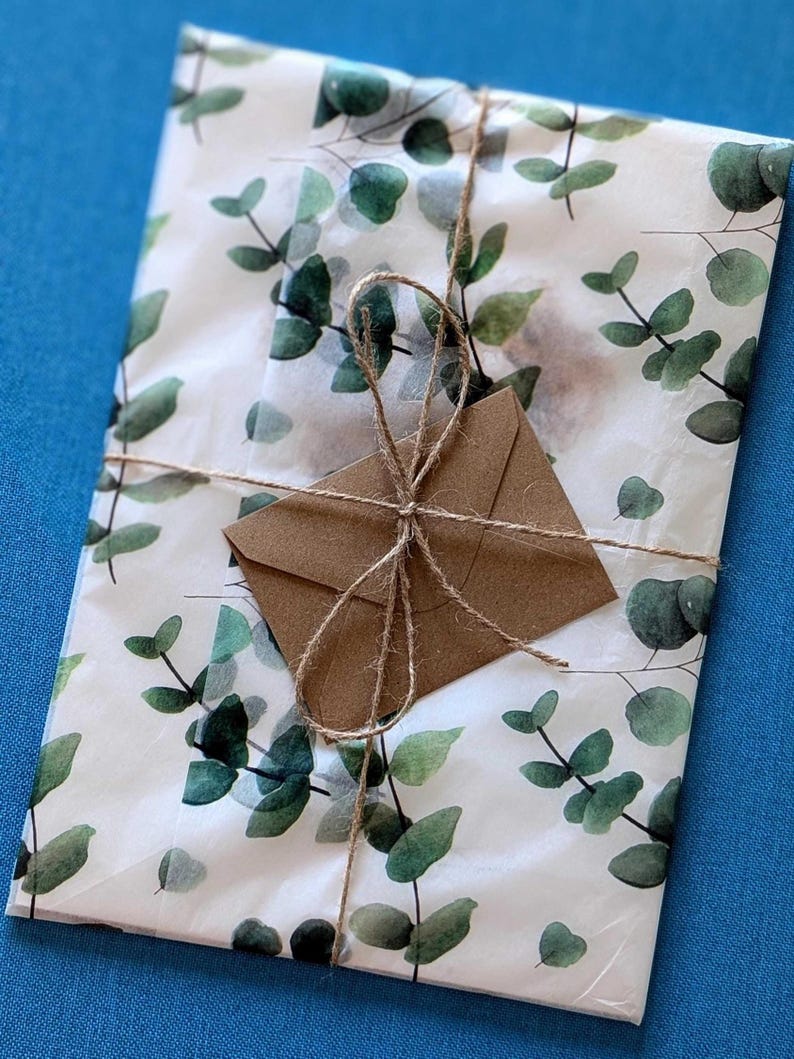 May include: A package wrapped in white paper with a green eucalyptus leaf pattern. The package is tied with twine and has a small brown envelope attached. The background is a solid blue.