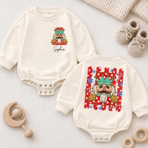 May include: Two cream-colored long-sleeve baby bodysuits with snap closures. One features a small nutcracker graphic and the name Sophia. The other displays a large nutcracker graphic surrounded by red stripes, candy canes, and blue snowflakes.