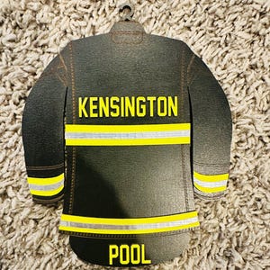 Może przedstawiać: Drewniana ozdoba w kształcie czarnej kurtki strażackiej. Kurtka ma żółte odblaskowe paski oraz żółte napisy "KENSINGTON" i "POOL". Na górze znajduje się mała pętla do zawieszenia.