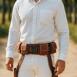 Può includere: Una cintura tattica in pelle marrone con più tasche e una fibbia in metallo. La cintura è indossata sopra una camicia e pantaloni bianchi. La cintura ha due fondine in pelle marrone.