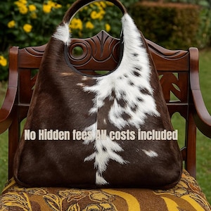 Op de afbeelding: Een grote schoudertas van bruin en wit koeienhuid met een enkele band. De tas staat op een houten stoel. De tekst "No Hidden fees All costs included" staat onderaan.