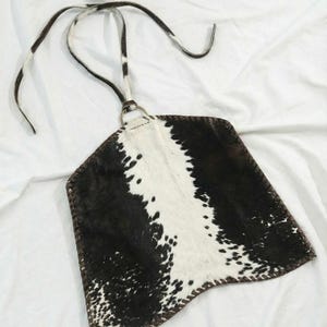 May include: A cowhide bag with a distinctive black and white spotted pattern. It features a brown leather trim and a neck strap. The bag is designed to be worn over the shoulder, offering a stylish accessory.