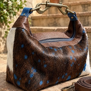 Può includere: Una borsa realizzata in pelle bovina marrone e bianca con pois blu. Presenta un pannello superiore in pelle marrone testurizzata con cerniera centrale, attacchi per tracolla blu e un'ampia maniglia metallica a forma di moschettone.