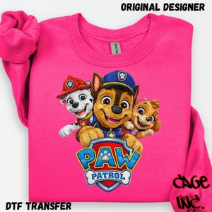 May include: A vibrant pink sweatshirt with a graphic of Paw Patrol characters Chase, Marshall, and Skye. The Paw Patrol logo is below the characters. The words "ORIGINAL DESIGNER" and "DTF TRANSFER" are printed on the sweatshirt.