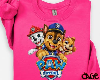 May include: A bright pink sweatshirt featuring a colorful graphic of the Paw Patrol characters: Chase, Marshall, and Skye. The Paw Patrol logo is displayed below the characters. The words "ORIGINAL DESIGNER" and "DTF TRANSFER" are printed on the sweatshirt.