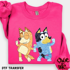 May include: A vibrant pink sweatshirt with a glittery graphic of two cartoon dogs. One dog is orange and the other is blue. The text "DTF TRANSFER" is at the bottom left.