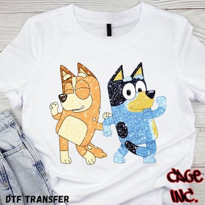 May include: White t-shirt featuring two cartoon dogs, one orange and one blue, with glitter effects. The orange dog is winking, and the blue dog is raising its arm. The text "DTF TRANSFER" is at the bottom left.