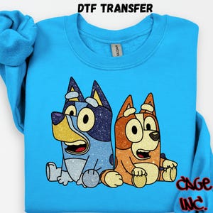 Puede incluir: Una sudadera azul claro con una transferencia DTF de dos perros de dibujos animados. Uno es azul con purpurina, el otro es naranja. El texto "DTF TRANSFER" está en la parte superior y "CAGE INC" en la inferior.