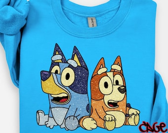 May include: A light blue sweatshirt featuring a DTF transfer of two cartoon dogs. One dog is blue with glitter, the other is orange. The text "DTF TRANSFER" is at the top, and "CAGE INC" is at the bottom.