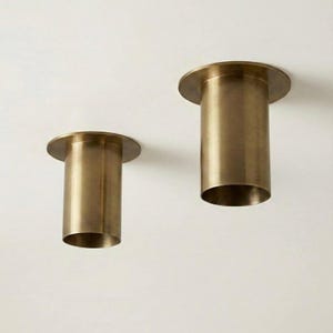May include: Two cylindrical, brass-colored light fixtures with a circular flange at the top. The fixtures are of different sizes, with one being shorter than the other. The open ends of the cylinders are visible, and they are set against a white background.