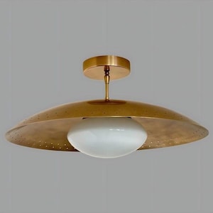 May include: A ceiling light fixture in brass. It features a wide, saucer-shaped shade with small perforations and a white, oval glass globe. The fixture is mounted on a brass stem and ceiling plate. The shade's diameter is approximately 50 cm.