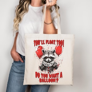 Könnte beinhalten: Cremefarbene Tragetasche mit einem Clown-Waschbär-Design, der ein Messer und rote Luftballons hält. Der Text lautet "YOU'LL FLOAT TOO!" und "DO YOU WANT A BALLOON?" in Rot. Halloween-Thema.