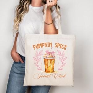Könnte beinhalten: Cremefarbene Tragetasche mit den Worten "Pumpkin Spice Social Club" in orangefarbener Schrift. Die Tasche zeigt eine Aquarellillustration eines Pumpkin Spice-Getränks mit einer rosa Schleife und einem rosa Blumenkranz.