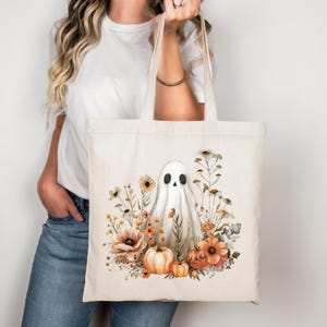 Könnte beinhalten: Cremefarbene Tragetasche mit einer Aquarellillustration eines Geistes, Kürbissen und Blumen in Orange, Gelb und Braun. Der Geist hat schwarze Augen und einen lächelnden Mund. Die Tasche hat zwei cremefarbene Griffe. Halloween-Thema.