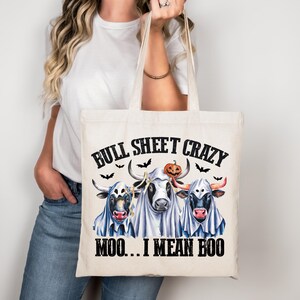 Può includere: Una borsa tote color crema con la scritta "BULL SHEET CRAZY" e "MOO... I MEAN BOO." La borsa presenta una grafica di tre mucche vestite da fantasmi, con una zucca e dei pipistrelli. La borsa ha manici color crema.