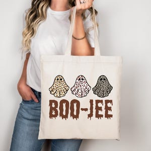 Könnte beinhalten: Eine cremefarbene Tragetasche mit drei Geisterillustrationen mit Leopardenmuster in Beige, Rosa und Schwarz. Der Text "BOO-JEE" ist in einer tropfenden braunen Schrift unter den Geistern aufgedruckt. Die Tasche hat zwei Henkel.