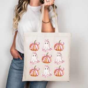 Op de afbeelding: Een beige draagtas met een herhalend patroon van aquarel pompoenen met roze strikken en vriendelijke spookillustraties. De tas wordt vastgehouden door een persoon die een wit t-shirt en een blauwe spijkerbroek draagt.