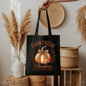 Könnte beinhalten: Schwarze Tragetasche mit einem Kürbismotiv und dem Text "FARM FRESH Pumpkins HAND PICKED DAILY". Der Kürbis ist orange mit einer Schleife mit Leopardenmuster. Die Tasche hat zwei schwarze Griffe.