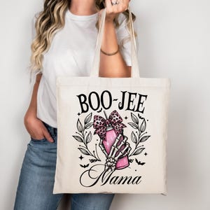 Może przedstawiać: Kremowa torba typu tote z czarnym napisem "BOO-JEE Mama". Projekt przedstawia różowy napój z kokardą w panterkę, trzymany przez szkieletową dłoń, otoczony liśćmi i nietoperzami. Torba ma długie uchwyty.