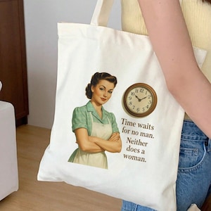 Könnte beinhalten: Cremefarbene Tragetasche mit einer Illustration im Retro-Stil einer Frau in grünem Kleid und Schürze, mit Uhr und dem Text "Time waits for no man. Neither does a woman."