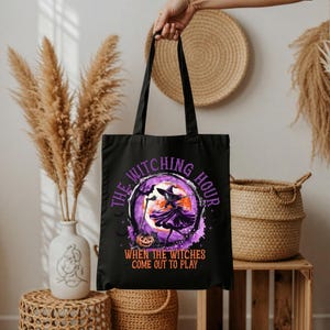 Può includere: Borsa tote nera con un design di Halloween. Il design include una silhouette di strega, una luna piena e il testo "THE WITCHING HOUR" e "WHEN THE WITCHES COME OUT TO PLAY". La borsa ha lunghi manici neri.