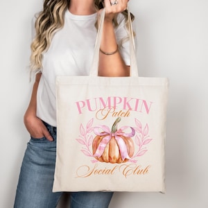 Könnte beinhalten: Beige Canvas-Tragetasche mit einem Aquarell-Kürbis-Design mit einer rosa Schleife und den Worten "Pumpkin Patch Social Club". Das Design ist von rosa Blättern umgeben. Die Tasche hat zwei Griffe.