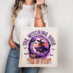 Può includere: Borsa tote color crema con un design a tema Halloween. La grafica include una strega, pipistrelli, una zucca e il testo "THE WITCHING HOUR" e "WHEN THE WITCHES COME OUT TO PLAY" in viola e arancione.