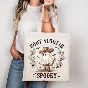 Könnte beinhalten: Eine cremefarbene Tragetasche mit dem Text "BOOT SCOOTIN' SPOOKY" und einem Cartoon-Geist mit Cowboyhut und -stiefeln. Das Design umfasst Fledermäuse und einen Kranz. Die Tasche hat zwei Griffe.