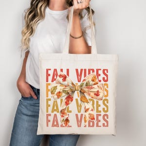 Könnte beinhalten: Cremefarbene Tragetasche mit den Worten "FALL VIBES" in Rot, Orange und Gelb. Eine große Schleife mit Herbstblättern ist mittig auf der Tasche. Die Tasche wird von einer Person gehalten.