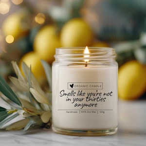 Könnte beinhalten: Eine brennende, weiße Kerze in einem durchsichtigen Glas. Die Kerze hat ein Etikett mit der Aufschrift "Smells like you're not in your thirties anymore". Die Kerze ist handgefertigt aus 100% Sojawachs und wiegt 120g. Zitronen und Grünzeug sind im Hintergrund.