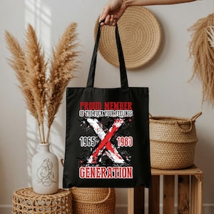 Op de afbeelding: Zwarte draagtas met de tekst "PROUD MEMBER OF THE FUCK YOUR FEELINGS GENERATION" in rood en wit. Het ontwerp bevat een grote witte "X" gekruist met een rode "X", en de jaren 1965 en 1980.