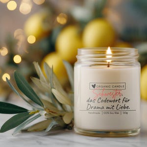 Pode incluir: Uma vela de cera de soja orgânica acesa em um frasco de vidro com o texto "Schwester, das Codewort für Drama mit Liebe." A vela é cercada por limões e vegetação.