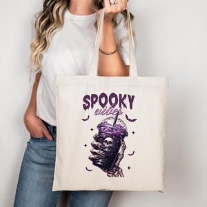 Może przedstawiać: Kremowa torba na zakupy z motywem Halloween. Grafika zawiera fioletowy napis "Spooky Vibes", fioletowy napój z czaszką i szkieletową dłoń. Czarne nietoperze i gwiazdy dopełniają projekt.