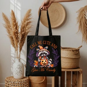 Könnte beinhalten: Schwarze Tragetasche mit Halloween-Motiv. Sie zeigt einen Waschbären mit Hexenhut, der einen Kürbis hält. Der Text "CALL ME CUTE AND GIVE ME CANDY" ist in Orange und Weiß gedruckt. Der Waschbär ist von lila Blättern umgeben.