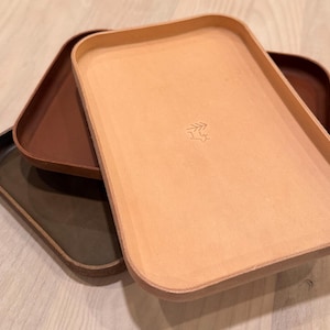 May include: Three rectangular trays in shades of brown, tan, and olive green are stacked. The top tray is light tan with a small leaf design. The trays have rounded corners and a smooth surface.