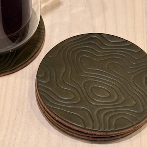 May include: A stack of olive green leather coasters with a topographic design. The coasters are round and have a brown edge. A glass with a dark liquid sits on one of the coasters.