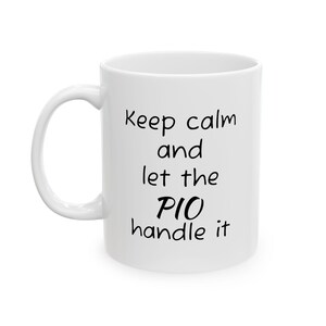 Könnte beinhalten: Weißer Keramikbecher mit gebogenem Henkel. Der Becher trägt den Schriftzug "Keep calm and let the PIO handle it" in schwarzer, handschriftlicher Schrift. Der Becher ist für Heißgetränke konzipiert.