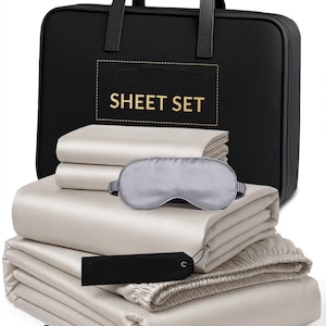 May include: A set of folded champagne-colored sheets, a matching sleep mask, and a black carrying case with the text SHEET SET printed in gold. The items are arranged with a black rectangular tag and several small green decorative beads.
