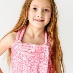 May include: A pink and white leopard print wrap towel with a halter neck design. The towel is wrapped around a person. The person has long, light brown hair and is smiling. The background is white.