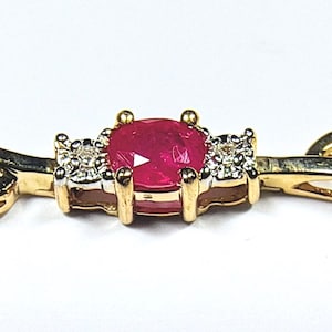May include: A gold bracelet featuring a central oval-cut red ruby gemstone flanked by two small clear accent stones on each side. The metal setting is yellow gold with a polished finish, and the band consists of open, curved gold links.